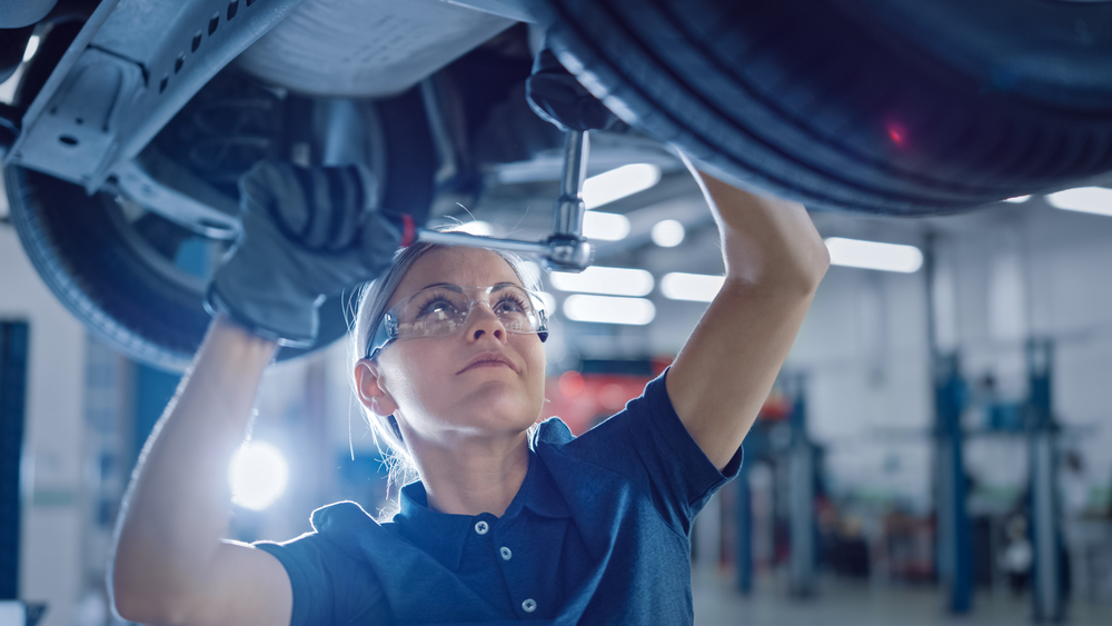 Kfz-Mechanikerin mit Schutzbrille repariert Auto auf Hebebühne